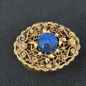 No label oval goldtone box with large blue stone in center.Tiny stones on top